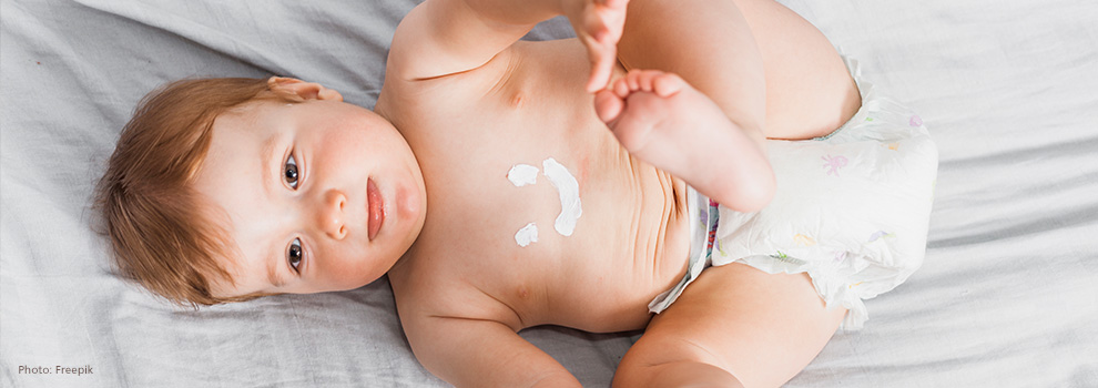 Baby liegt in Windel auf dem Rücken, Arme und Beine oben, Bauch mit Cremegesicht bemalt.