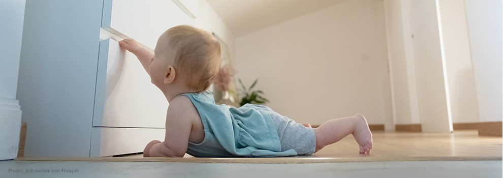 Kleinkind liegt auf dem Boden, streckt sich nach oben und versucht eine Schublade zu öffnen