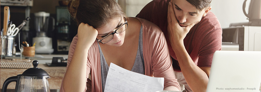Nachdenkliche Mutter sitzt am Tisch vor einem Laptop und hält Elterngeldantrag in den Händen, der Vater steht dabei ist am grübeln.