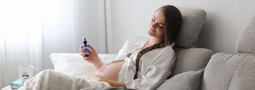 werdende Mutter sitzt mit freien Babybauch auf der Couch und hält Motherlove Geburts- und Babymassageöl in der Hand