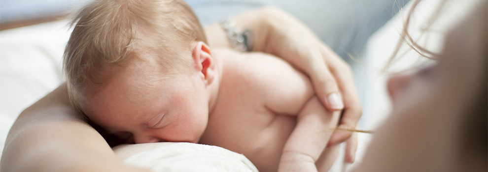 Ein Baby trinkt mit geschlossenen Augen an der Brust, das Gesicht der Mutter ist halb sichtbar.