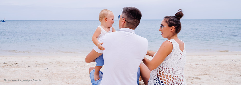 Eltern sitzen eng am Strand, das Kleinkind steht auf Papas Beinen und schaut zur Mutter.