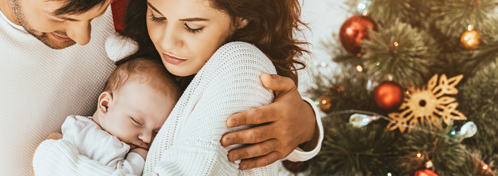 Glücklich Eltern mit schlafenden Baby im Arm stehen vor einem Weihnachtsbaum
