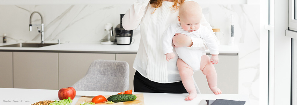 Mutter steht in der Küche und hält ihr Baby auf dem Arm. Auf dem Tisch vor hier liegt etwas Gemüse auf einem Schneidebrett