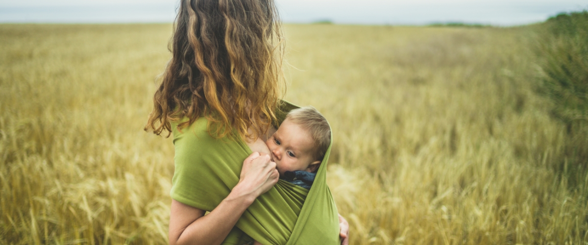Mutter steht in einem Kornfeld und stillt ihr Baby im Tragetuch