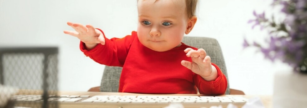 Kleinkind sitzt am Schreibtisch vor einer Tastatur und hebt die Hände, als ob schreiben möchte