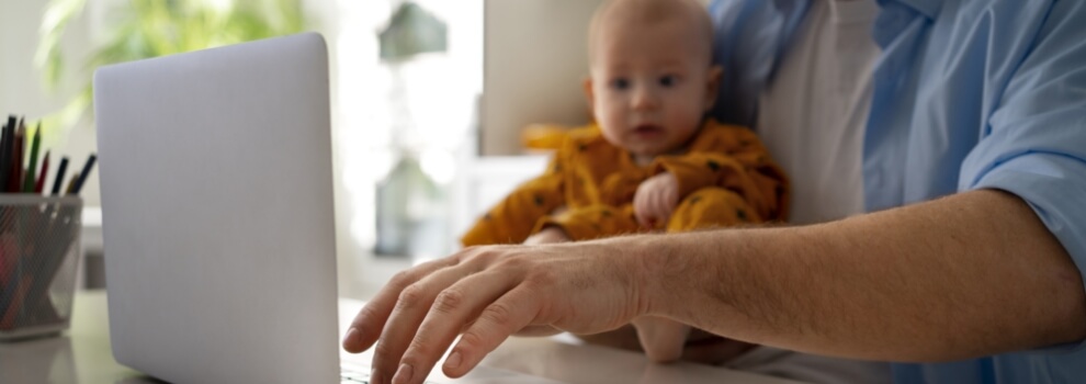 Vater sitzt vor dem aufgeklappten Laptop und das Baby auf seinem Schoß schaut in den Bildschirm