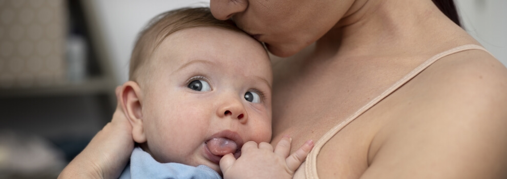 Mutter halt ihr Baby eng im Arm und küsst es auf den Kopf, Baby streckt die Zunge raus